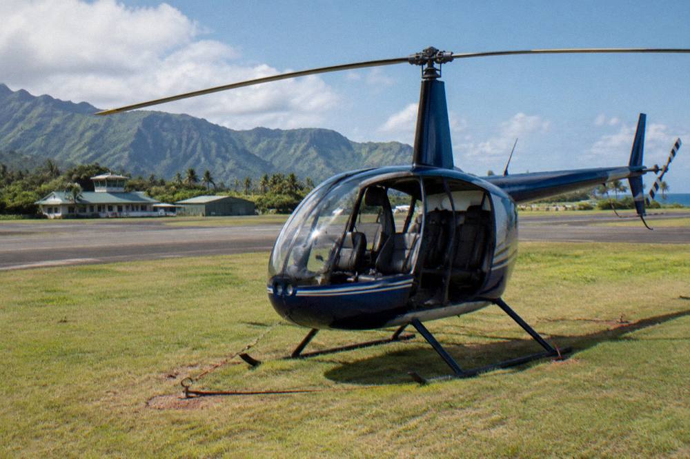 A small four-seat helicopter with its doors off parked on a grass pad near a Kauai heliport